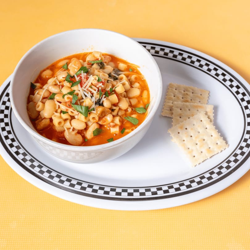 A white bowl of pasta fagioli soup with crackers on a checkered plate.