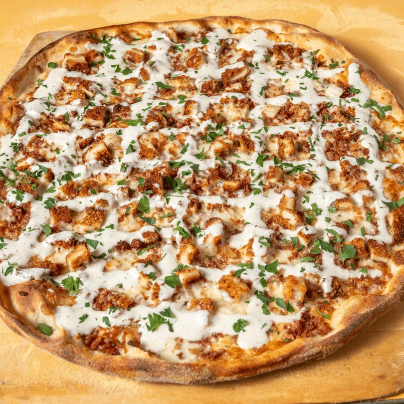 A whole pizza topped with crispy chicken, white sauce, and fresh parsley on a wooden peel.