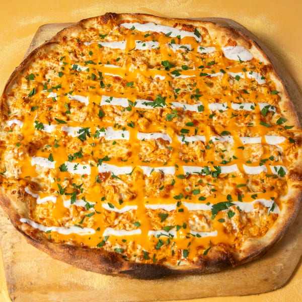 Buffalo chicken pizza with orange sauce, white ranch drizzle, and green parsley on a wooden peel.