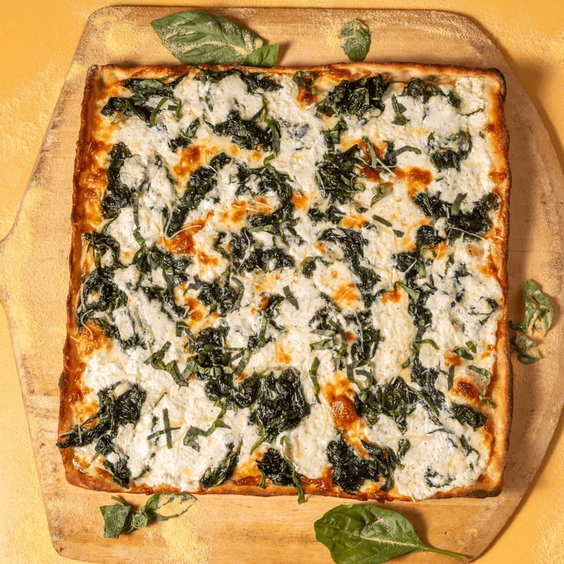 Square white pizza with spinach and ricotta cheese, garnished with fresh basil on a wooden peel.