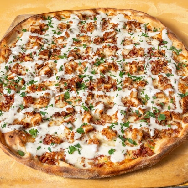 A whole pizza topped with crispy chicken, white sauce, and fresh parsley on a wooden peel.