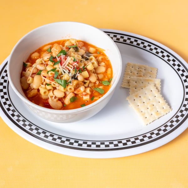 A white bowl of pasta e fagioli soup with crackers on a checkered plate.
